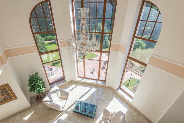 This image showcases the interior of a professionally photographed vacation rental property. The open-concept space includes double-height ceilings with exposed wooden beams, skylights providing natural light, a decorative iron balcony railing on the mezzanine level, an exposed brick accent column, and warm tile flooring throughout. The layout flows into a kitchen and dining area in the background, demonstrating the type of high-quality listing photography recommended with at least 30 high-resolution images sequenced from exterior to living spaces to bedrooms and amenities